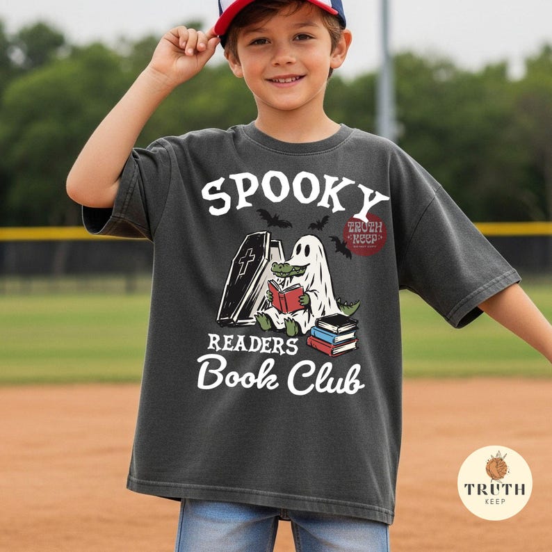 May include: A charcoal gray t-shirt featuring a graphic of a green alligator dressed as a ghost, sitting next to a coffin and reading a red book. The text reads "SPOOKY READERS Book Club" in white, with a small stack of books and bats.