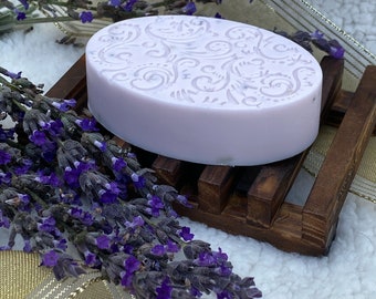 Könnte beinhalten: Eine Lavendel-Seife mit einem dekorativen Wirbelmuster, die auf einem Holzseifenhalter liegt. Die Seife ist helllila und hat ein natürliches, erdiges Aussehen.