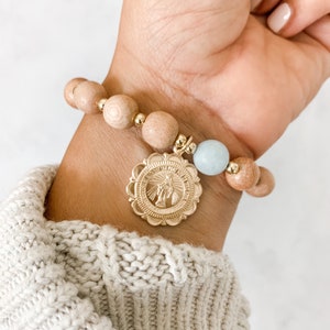 May include: A wooden beaded bracelet with a gold charm featuring a religious figure. The bracelet also includes a light blue stone bead.