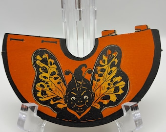 May include: An orange Halloween decoration featuring a black and yellow butterfly-like creature. The vintage-style image is mounted on a clear plastic display stand. The creature has a smiling face and is set against a black border.