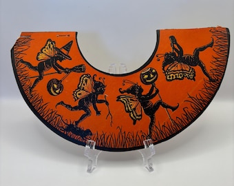May include: Orange Halloween decoration with black illustrations of fairies, a witch, and jack-o'-lanterns. The decoration has a curved shape with a black border and is propped up on a clear stand. The design is vintage and festive.