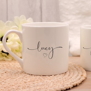 May include: Two white ceramic mugs with curved handles. One mug has the name "lucy" and a heart symbol, the other has "james" and a heart. The mugs are on a woven mat.