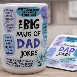 May include: A white ceramic mug with the text "THE BIG MUG OF DAD JOKES" in black and blue. The mug is covered in dad jokes in speech bubbles. A matching coaster with more jokes is next to the mug.