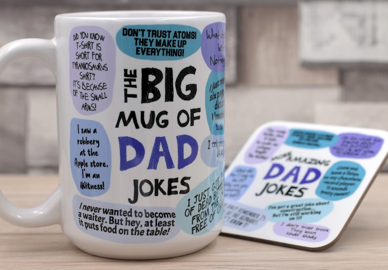 May include: A white ceramic mug with the text "THE BIG MUG OF DAD JOKES" in black and blue. The mug is covered in dad jokes in speech bubbles. A matching coaster with more jokes is next to the mug.