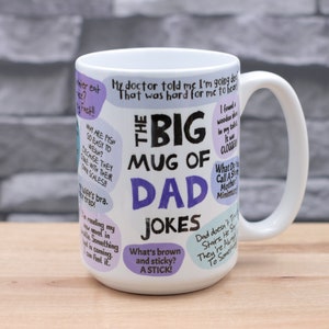 May include: A white ceramic mug with a large handle, featuring the text "THE BIG MUG OF DAD JOKES" in black and purple. The mug is covered in various dad jokes in speech bubbles. The mug is on a wooden surface.