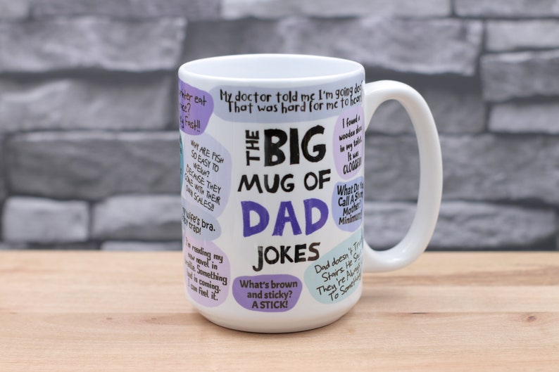 May include: A white ceramic mug with a large handle, featuring the text "THE BIG MUG OF DAD JOKES" in black and purple. The mug is covered in various dad jokes in speech bubbles. The mug is on a wooden surface.