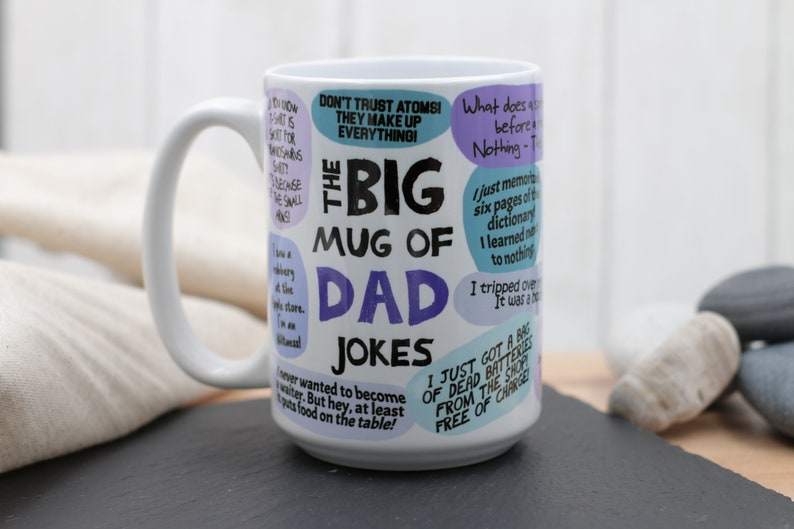 May include: A white ceramic mug with a large handle, featuring the text "The Big Mug of Dad Jokes" in black and purple. The mug is covered in various jokes in speech bubbles. The mug is on a black surface.