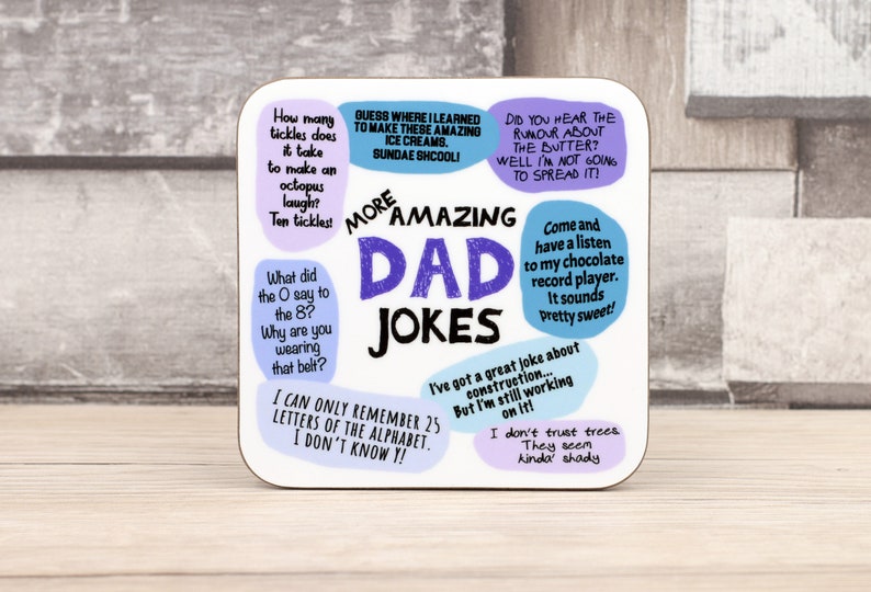 May include: A square coaster with the words "More Amazing Dad Jokes" in a playful font. The coaster features various jokes in speech bubbles, with a light background and colorful text.