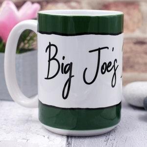 May include: White ceramic mug with a green band around the top and bottom. The mug has black text that says "Big Joe's" in a cursive font.