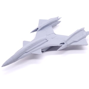 May include: A light gray model airplane, possibly a fighter jet, is positioned on a white background. The model has a sleek design with wings and tail fins. The plane's surface has visible textures.