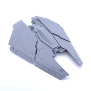 May include: A gray, futuristic-looking model of a spacecraft or vehicle. The model has a detailed, angular design with wings and a central cockpit area. The object is made of a solid material and is set against a white background.