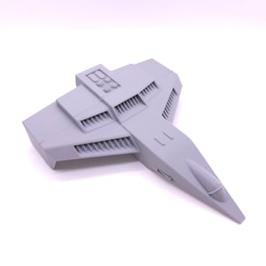 May include: A gray 3D printed model of a futuristic spaceship with a pointed nose and a large wingspan. The model has a detailed design with multiple air intakes and vents.