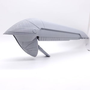 以下が含まれることがあります： 未来的な航空機または宇宙船のコンポーネントの、薄いグレーの3Dプリントモデル。デザインは、尖ったフロント、翼のような構造、角度の付いたフィンが特徴です。このモデルはおそらくプロトタイプです。