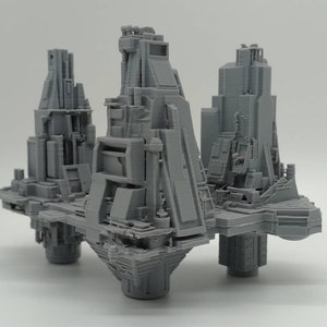 Può includere: Un modello stampato in 3D di una città futuristica con tre torri collegate da una piattaforma. Le torri sono in plastica grigia e hanno una varietà di forme geometriche.