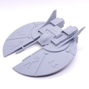 May include: A gray 3D printed model of a spaceship with a disc-shaped body and two wings. The spaceship has a detailed design with various lines and markings.