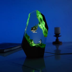 May include: A green resin lamp with a black base. The lamp features a small figurine of a scarecrow and a flying object inside the resin. The lamp is lit up with a green glow.