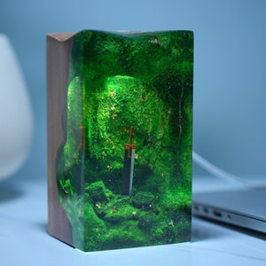 May include: A wooden box with a clear resin top containing a miniature sword and green moss. The moss is a vibrant green and the sword is black with a gold hilt.