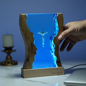 May include: A wooden base with a blue resin insert featuring a silver crucifix. The resin is illuminated with blue LED lights. The base has the text "RESIN ART YPC" printed on it.