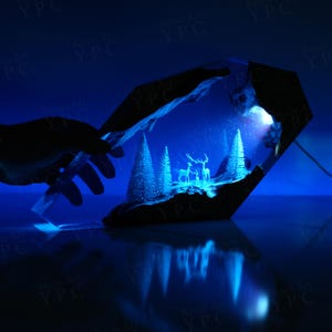 May include: A resin art piece with a winter scene inside. The scene features a snowy forest with two deer and a glowing light source. The resin is clear and the scene is visible from all sides.