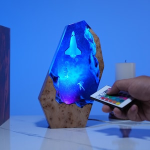 Puede incluir: Lámpara decorativa de resina con temática espacial, con una lanzadera y astronautas en una galaxia azul y morada. La lámpara tiene una base de madera y se controla con un mando a distancia.