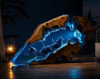 Op de afbeelding: Een decoratieve lamp gemaakt van natuurlijk hout en doorschijnende blauwe hars. Het interieur bevat een gedetailleerd miniatuur scheepswrak en een kwallenfiguurtje verlicht door een felblauw licht, wat een onderwaterscène in het hout creëert.