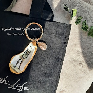 May include: A gold keychain with an oyster charm featuring a painted image of a person. The keychain is attached to a black tote bag with the text "Slow Boat Studio" and "oh life" printed on it.