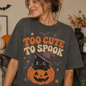 Pode incluir: Uma t-shirt de manga curta na cor pimenta com um gráfico de um gato preto usando um chapéu de bruxa roxo, sentado dentro de uma abóbora laranja. O texto "TOO CUTE TO SPOOK" está impresso em letras laranja e creme acima da ilustração.