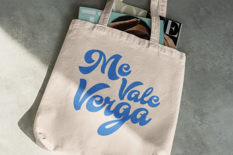 以下が含まれることがあります： ベージュのトートバッグ。青い筆記体で「Me Vale Verga」と書かれています。バッグの中には雑誌が入っています。丈夫な生地で作られており、持ち運び用のハンドルが2つ付いています。
