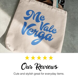 以下が含まれることがあります： クリーム色のトートバッグ。青い筆記体で「Me Vale Verga」と書かれています。丈夫な生地で作られており、シンプルなデザインです。バッグの中には雑誌が見えます。