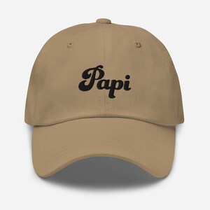 May include: A khaki dad hat with black embroidered text that says "Papi" in a retro font.