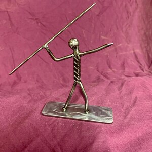 May include: A metal sculpture of a person throwing a javelin. The sculpture is made of twisted metal and is standing on a metal base.