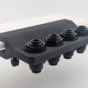 May include: A black 3D-printed clamp with four black fittings. The clamp is attached to a white rectangular object. The clamp has a screw for tightening. The fittings have a hexagonal shape.