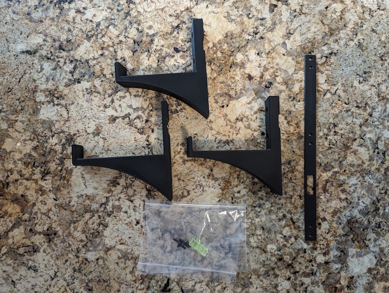 May include: A set of four black metal shelf brackets with a curved design. The brackets are packaged with screws in a clear plastic bag. A long black metal strip with holes is also included.