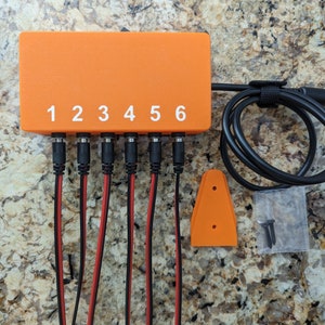May include: Orange power supply box with six numbered ports and black cables. The box is labeled with numbers 1 through 6. The power supply box is on a brown and white speckled surface.