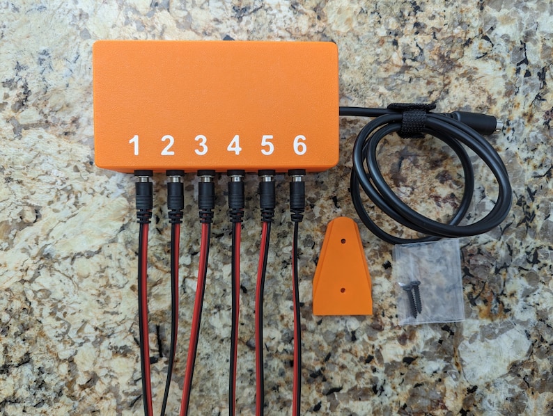 May include: Orange power supply box with six numbered ports and black cables. The box is labeled with numbers 1 through 6. The power supply box is on a brown and white speckled surface.