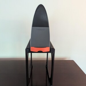 May include: A gray and orange device with the word "TRIDENT" on it, sitting on a black stand. The stand has two legs and is on a dark brown surface.
