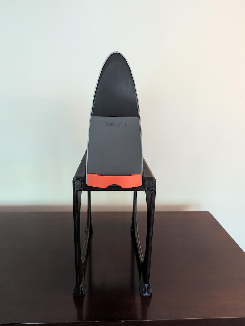 May include: A gray and orange device with the word "TRIDENT" on it, sitting on a black stand. The stand has two legs and is on a dark brown surface.