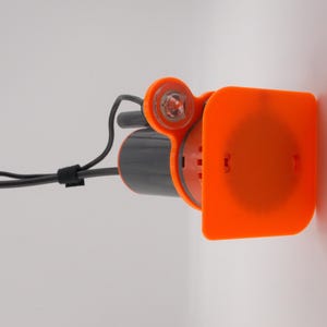 Pu&ograve; includere: Un dispositivo arancione e grigio con una luce e un cavo nero. Il dispositivo ha una copertura rettangolare arancione e una luce rotonda arancione. Il cavo &egrave; fissato con una cinghia nera.