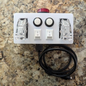 May include: A white plastic panel with two clear toggle switches, two round white buttons, and two red buttons. The panel is connected to a black USB cable.