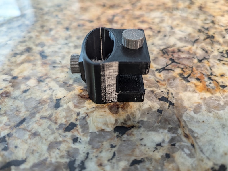 May include: A black 3D printed camera mount with a gray knob. The mount has a circular opening and a slot for attaching a camera.