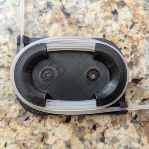 May include: A black 3D printed holder with two circular openings for holding tubing. The holder has a white tube wrapped around it.