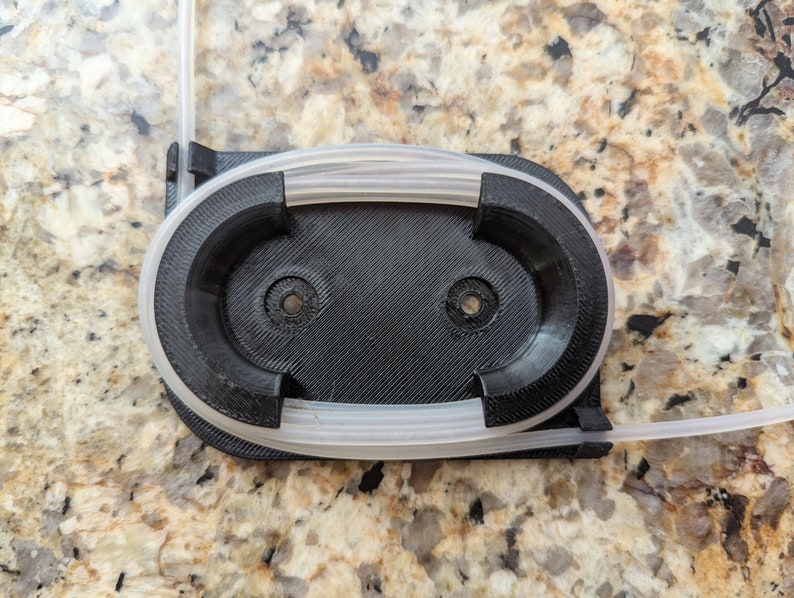 May include: A black 3D printed holder with two circular openings for holding tubing. The holder has a white tube wrapped around it.