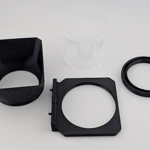 May include: An assortment of black and clear camera accessories, including a lens hood, a filter holder, a circular ring, and a clear glass component. The items are arranged on a white surface.