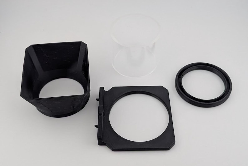 May include: An assortment of black and clear camera accessories, including a lens hood, a filter holder, a circular ring, and a clear glass component. The items are arranged on a white surface.