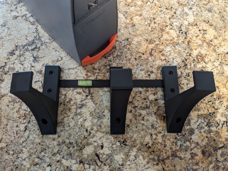 May include: Black metal wall mount with three hooks, each with a curved design. The hooks are attached to a metal bar with screws. A level is placed on the bar.