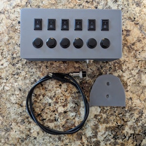 May include: A grey box with six black toggle switches and six black push buttons. The box is connected to a black cable with a connector on each end. There is also a grey plastic piece with two holes in it.