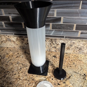 May include: A 3D-printed, black and white kitchen tool set. The set includes a tall, cylindrical container with a flared top, a small hexagonal rod on a circular base, and a clear, circular lid with a central protrusion. The items are on a speckled countertop.