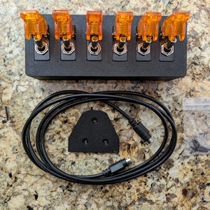 Puede incluir: Una caja negra con seis interruptores de palanca naranja, un cable negro con un conector, una almohadilla de espuma negra con tres agujeros y una peque&ntilde;a bolsa de pl&aacute;stico con tornillos.