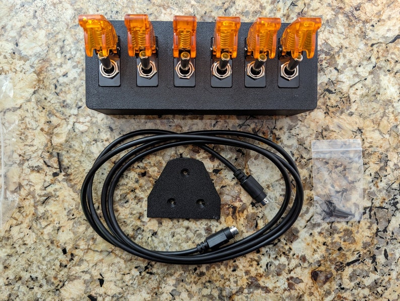 Puede incluir: Una caja negra con seis interruptores de palanca naranja, un cable negro con un conector, una almohadilla de espuma negra con tres agujeros y una peque&ntilde;a bolsa de pl&aacute;stico con tornillos.