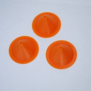 Pu&ograve; includere: Tre imbuti di plastica arancione con un piccolo foro sulla punta. Gli imbuti sono su una superficie bianca.
